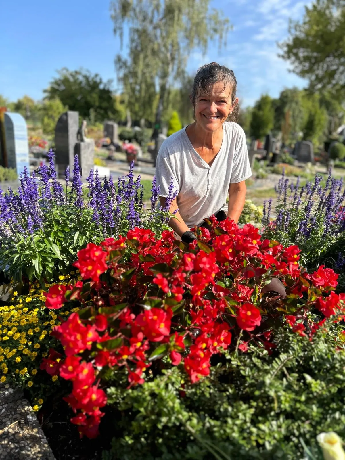 Waltraud Zenth kniet lächelnd hinter einem Beet mit roten Begonien und blauem Salbei auf dem Friedhof